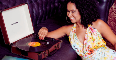 Carolina Frade sorrindo enquanto coloca um disco de vinil para tocar em uma vitrola portátil.